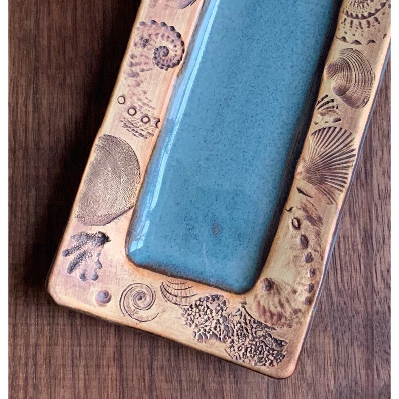 May include: A rectangular, handcrafted ceramic tray with a teal center and a textured, golden-brown border. The border is adorned with various seashell and spiral designs, creating a coastal theme.