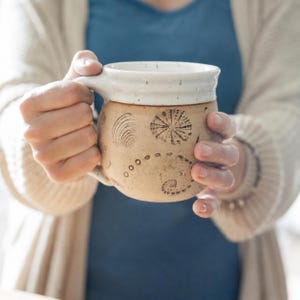 Könnte beinhalten: Eine beige Keramiktasse mit weißem Rand und Henkel. Die Tasse hat geätzte Muschelmotive und eine Reihe kleiner Punkte. Die Tasse wird von einer Person gehalten, die ein blaues Hemd und eine cremefarbene Strickjacke trägt.