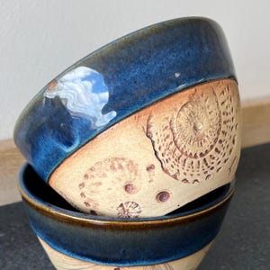 May include: Two ceramic bowls stacked. The top bowl has a glossy, dark blue exterior and a tan interior with a spiral seashell design. The bottom bowl is also dark blue with a tan interior and seashell design.