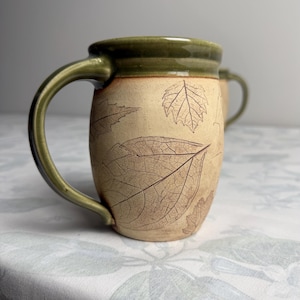 Puede incluir: Una taza de cerámica con cuerpo beige que presenta patrones de hojas en relieve de color marrón oscuro y un esmalte verde oliva liso en el borde y el asa. La taza tiene una forma redondeada y un asa lateral resistente, con otra igual detrás.