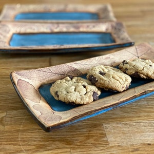 May include: Rectangular ceramic serving dish with a blue center and leaf pattern. The dish holds three chocolate chip cookies. The dish has a brown and blue color scheme.