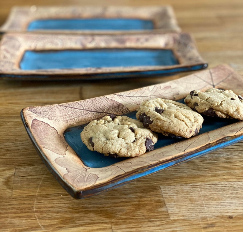 May include: Rectangular ceramic serving dish with a blue center and leaf pattern. The dish holds three chocolate chip cookies. The dish has a brown and blue color scheme.