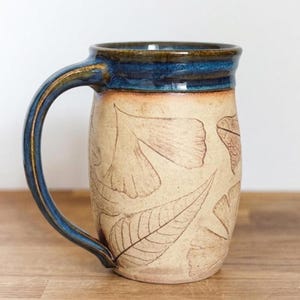 May include: A ceramic mug with a cream-colored body featuring embossed leaf patterns. The rim and the curved handle are finished in a deep, glossy blue glaze that contrasts with the textured, matte surface of the main body of the vessel.