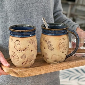 May include: Two ceramic mugs with a beige base and dark blue rim and handle. The mugs are decorated with seashell imprints. One mug has a spoon inside. The mugs are on a wooden tray.