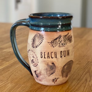 May include: A ceramic mug with a light brown body and a teal blue rim and handle. The mug has a design of seashells and the text "Beach Bum" on the front.