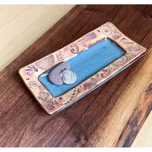 May include: A rectangular, handcrafted dish with a light blue interior and a copper-colored, seashell-patterned border. A small, gray stone and a white seashell are placed inside the dish. The dish is displayed on a dark wooden surface.