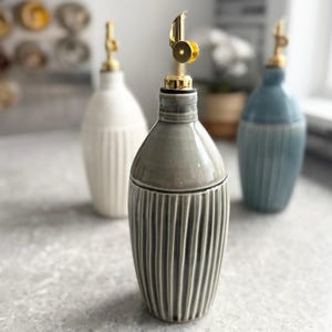 May include: Three ceramic oil dispensers with a ribbed texture and a glossy finish. Each bottle features a gold-colored metal pour spout. The dispensers are colored in shades of grey, white, and blue, standing upright in a group on a flat surface.