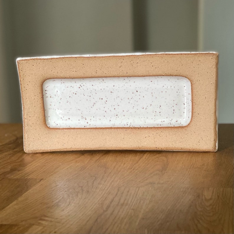 May include: A rectangular ceramic serving dish with a speckled white interior and a light brown exterior. The dish has a raised, rectangular interior section. The exterior has a slightly rough texture, and the dish sits on a wooden surface.