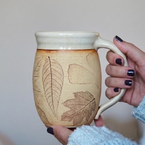 May include: A beige ceramic mug with a cream-colored rim and handle. The mug features detailed brown leaf imprints. The mug is held by a person wearing a light blue sweater.