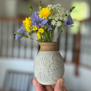 May include: A small, handmade ceramic vase with a light gray body and a brown rim, filled with a bouquet of wildflowers. The vase features a carved floral design. The flowers include yellow, blue, and white blossoms.