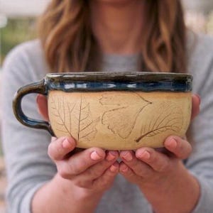 May include: A ceramic mug with a wide, shallow bowl shape and a single handle. The exterior features a natural tan, unglazed finish with embossed leaf patterns, while the rim and handle are finished in a glossy, dark navy blue glaze.