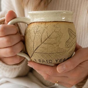 May include: A round ceramic mug with a speckled cream-colored rim and a tan body featuring embossed leaf patterns. The text I SAID YES is imprinted in black lettering near the base of the mug. The mug has a curved handle on the left side.