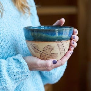 May include: A handmade ceramic bowl with a blue glazed rim and a beige lower section. The beige section features a detailed leaf design. The bowl is held in hands, suggesting its size and use.