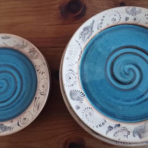 May include: Two ceramic plates with a teal blue spiral design in the center. The plates have a cream-colored rim with seashell and spiral patterns. The plates are on a wooden surface.