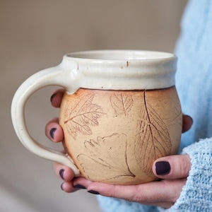 May include: A handmade ceramic mug with a cream-colored rim and handle. The body of the mug is tan with embossed leaf designs. The mug is held in a person's hands, with a light blue sweater in the background.