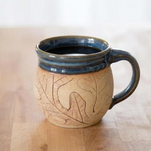 May include: A ceramic mug with a rounded body and a curved handle. The upper rim and handle are glazed in a deep blue, while the lower portion features a light tan, unglazed surface decorated with embossed leaf patterns across the exterior.