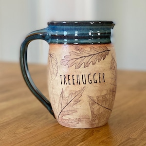 Könnte beinhalten: Ein Keramikbecher mit glänzend blauem Rand und Henkel, dessen hellbeiger Körper mit dunkelbraunen Blattabdrücken verziert ist. Das Wort TREEHUGGER ist in schwarzen Großbuchstaben auf der Vorderseite aufgedruckt, umgeben von verschiedenen Blattmustern.