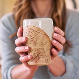 May include: A handmade ceramic mug with a beige glaze and leaf imprints. The lower portion of the mug features detailed leaf patterns in a natural brown color. The top half is a lighter beige. The mug is held by a person.