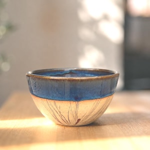 May include: A handmade ceramic bowl with a two-tone design. The top half is a deep blue glaze, while the bottom half is a cream color with delicate, etched lines. The bowl sits on a wooden surface.