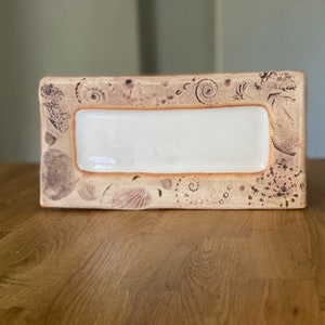 May include: Rectangular ceramic dish with a cream-colored center and a tan border. The border is decorated with seashell and spiral designs in a darker brown color. The dish is placed on a wooden surface.