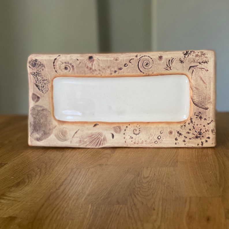 May include: Rectangular ceramic dish with a cream-colored center and a tan border. The border is decorated with seashell and spiral designs in a darker brown color. The dish is placed on a wooden surface.