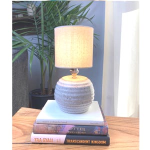 May include: A ceramic table lamp with a light beige fabric shade sits on a stack of books. The lamp has a round, light blue and grey base. The books are titled "Everyday Sacred", "Harry Potter and the Sorcerer's Stone", and "Transcendent Kingdom".