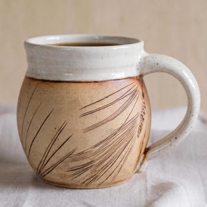 Op de afbeelding: Een handgemaakte keramische mok met een beige body en een witte rand. De mok heeft een geëtst ontwerp van tarwearen. De handgreep en de rand zijn gespikkeld. De mok is gevuld met een donkere vloeistof.
