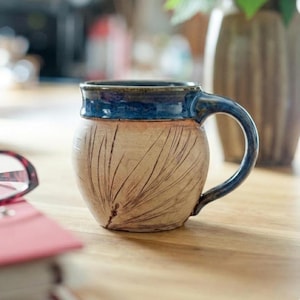 Może przedstawiać: Ręcznie wykonany ceramiczny kubek z niebieską obwódką i uchwytem. Kubek ma beżowy korpus z wytrawionymi liniami i zaokrąglonym kształtem. Kubek stoi na drewnianej powierzchni.
