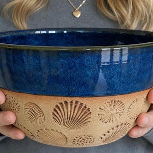 May include: A handcrafted ceramic bowl with a deep blue glazed interior and a tan exterior. The exterior features embossed seashell designs. The bowl is held by a person, showcasing its size and shape. This decorative bowl is suitable for serving or display.