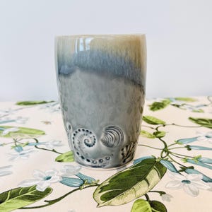 May include: A cylindrical ceramic tumbler featuring a smooth, glossy glaze that transitions from a soft beige at the top to a deep, muted blue-grey at the base. The lower section is decorated with embossed spiral and seashell motifs.