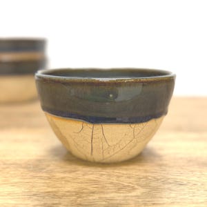 Puede incluir: Un cuenco pequeño de cerámica con un borde esmaltado en azul oscuro que transiciona hacia una base de color beige claro. La parte inferior del cuenco muestra una textura de hoja impresa con detalles de grietas en la superficie.