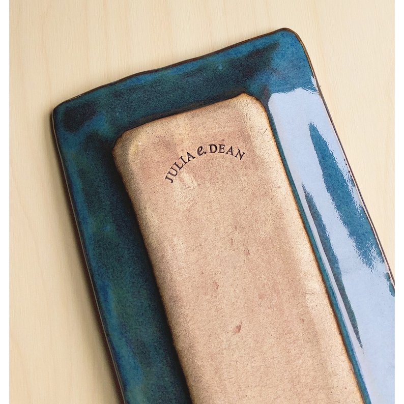 May include: Two rectangular ceramic serving platters, one nested inside the other. The top platter has a glossy, deep teal glaze with a slightly irregular edge. The bottom platter is a lighter tan color with the text "JULIA e. DEAN" printed on it.