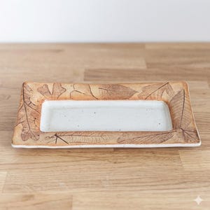 May include: Rectangular ceramic serving dish with a white interior and a brown leaf-patterned border. The dish is set on a wooden surface. The leaves are detailed and add a natural, rustic aesthetic.