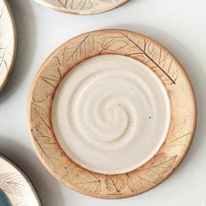 May include: A round ceramic plate featuring a smooth, cream-colored center with a subtle spiral texture. The wide, tan-colored rim is decorated with detailed, dark brown leaf imprints that wrap around the entire circumference of the plate.
