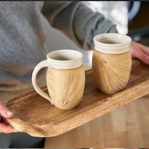 May include: Two beige ceramic mugs with a cream-colored rim and handle, and a leaf design. The mugs are on a wooden tray. The mugs are handmade and have a rustic, natural look. The mugs are perfect for coffee or tea.