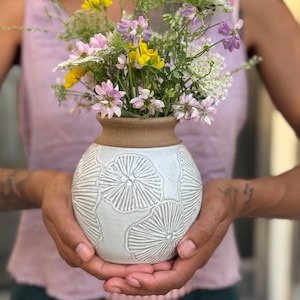May include: A white ceramic vase with a textured floral pattern, holding a bouquet of colorful wildflowers. The vase has a brown rim and is held in both hands.