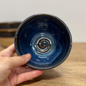May include: A round ceramic bowl with a deep blue glazed interior featuring a prominent brown spiral design at the center. The rim of the bowl is finished with a dark brown edge, and the overall surface has a smooth, glossy texture throughout.