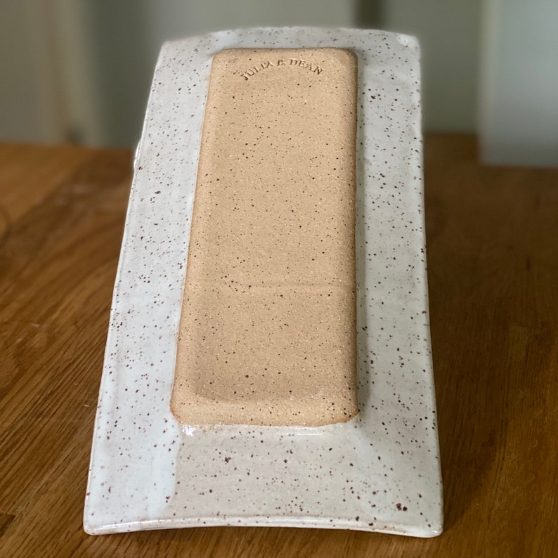 May include: Rectangular ceramic serving platter with a speckled white border and a light brown center. The words "Julia E. Dean" are stamped into the center. The platter is on a wooden surface.