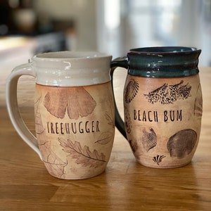 Puede incluir: Dos tazas de cerámica con diseños diferentes. La taza de la izquierda tiene un esmalte blanco y un diseño marrón de hojas con el texto "TREEHUGGER". La taza de la derecha tiene un esmalte azul y un diseño marrón de conchas marinas con el texto "BEACH BUM".