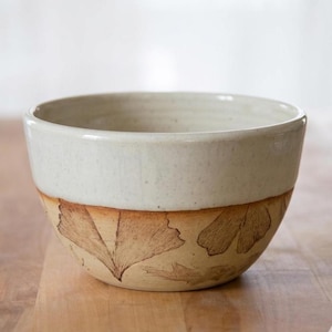May include: A handmade ceramic bowl with a cream-colored upper section and a lower section featuring a leaf design in shades of brown. The bowl is round and appears to be suitable for serving food or as a decorative piece.