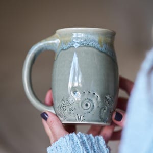 May include: A ceramic mug with a glossy sage green glaze and a curved handle. The lower portion of the mug features embossed spiral and circular patterns, while the upper rim displays a subtle drip effect with lighter blue and tan tones.