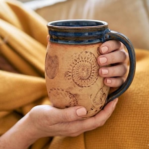 Könnte beinhalten: Eine handgefertigte Keramik-Tasse mit beigefarbenem Äußeren und dunkelblauem Innenraum und Henkel. Die Tasse hat geprägte Spiral- und Fingerabdruckmuster. Der Rand der Tasse ist mit einem dunkelblauen Band akzentuiert.