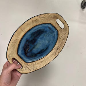 May include: An oval ceramic serving tray with a deep blue, glossy glazed center and a wide, tan-colored rim featuring etched leaf patterns. The tray includes two integrated handles on opposite ends, with a dark, contrasting edge finish.