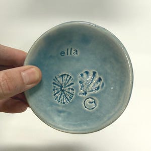 May include: A round, light blue ceramic trinket dish with a glossy finish. The interior features the stamped name "ella" at the top, alongside three distinct embossed botanical impressions including a circular leaf, a jagged leaf, and a small spiral.