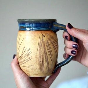Könnte beinhalten: Eine handgefertigte Keramiktasse mit blauem Henkel und Rand. Die Tasse hat einen beigefarbenen Boden mit braunen geätzten Linien und wird von einer Person gehalten. Die Tasse ist ein einzigartiges Keramikstück.