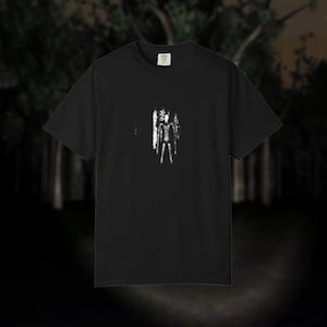 Op de afbeelding: Een zwart t-shirt met korte mouwen en een kleine, gecentreerde witte afbeelding van een lange, slanke, gezichtsloze figuur met lange armen. Het ontwerp heeft een verweerde, getextureerde uitstraling. Het shirt heeft een ronde hals.