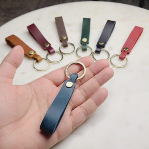 May include: A collection of leather keychains in various colors, including brown, burgundy, green, navy, and red. Each keychain features a leather strap attached to a brass ring with a brass rivet.