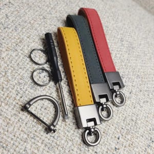 Op de afbeelding: Leren sleutelhangers in geel, donkergroen en rood. Elke sleutelhanger heeft een metalen sluiting en een lus voor sleutels. Een kleine schroevendraaier, twee sleutelringen en een metalen D-ring zijn ook aanwezig.