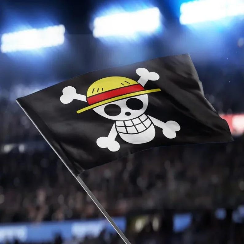 May include: Black flag featuring a white skull and crossbones design with a yellow hat and red band. The flag is waving in front of a blurred background, possibly a stadium or arena. The flag is held by a white pole.