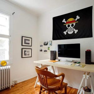 May include: A black flag with a white skull and crossbones design, wearing a yellow and red straw hat, hangs on a white wall. The flag is above a wooden desk with a computer, speakers, and a wooden chair. The room has a window and hardwood floors.